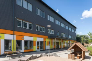 Nedre våningsplanet där förskolan finns har en fasad i lekfulla färger i orange, gult och vitt.  I förgrunden syns en sandlåda och en lekstuga. De två övre våningarna är mörkt gråa.