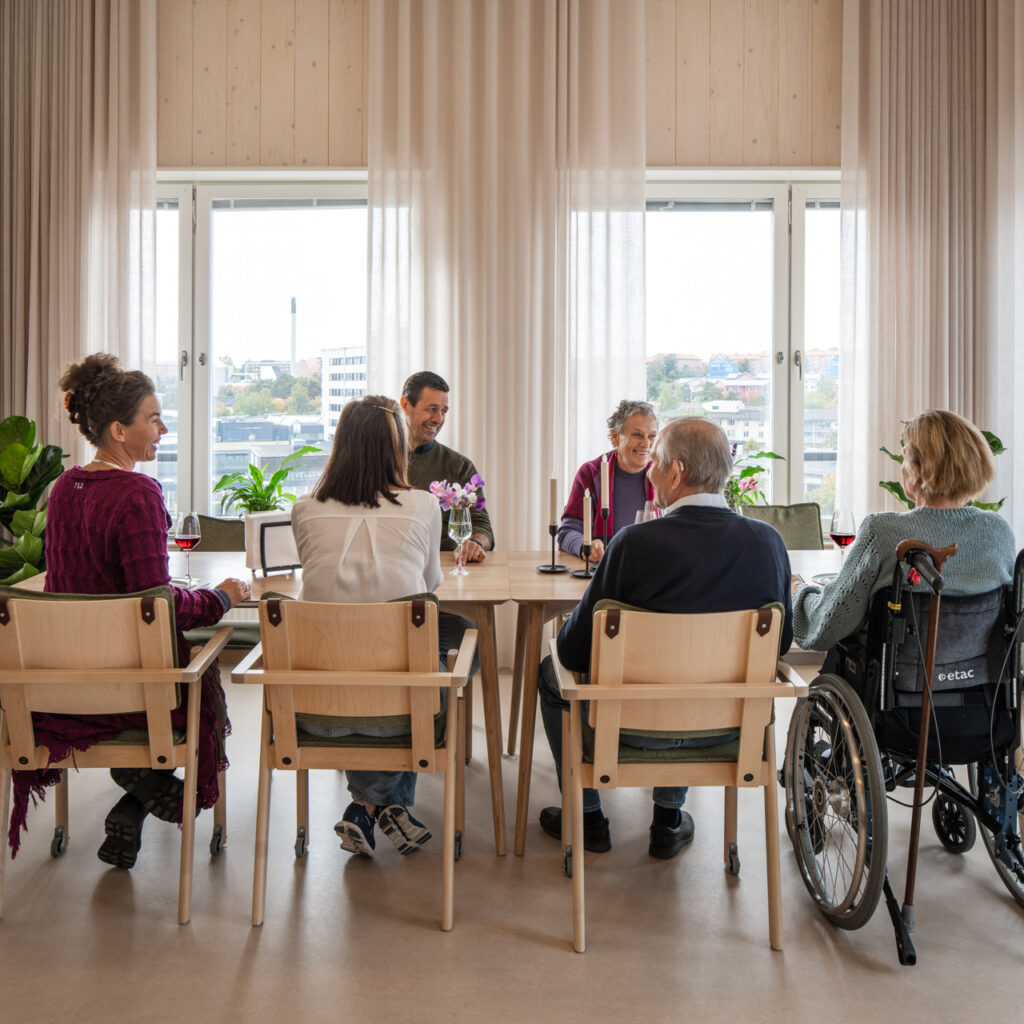 A small gathering of people sitting around a table at a nursing home and are having a pleasant conversation.